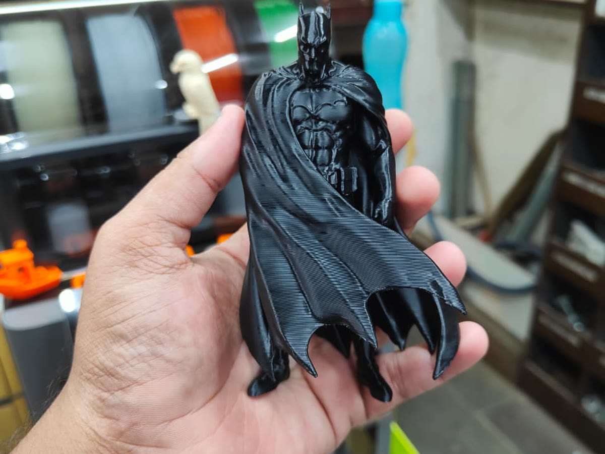 A printed Batman figure held in a hand, filament spools and the printer behind it.